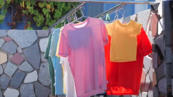 Na het wassen drogen het ondergoed en de kleding na het wassen op de waslijn, T-shirts, roze kleren, gele, felle kleuren op de waslijn buiten bij zonnig weer. — Stockvideo