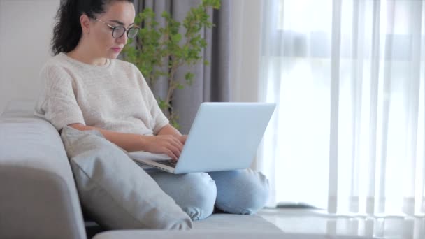 Ung seriös kvinna frilansare arbetar på en bärbar dator sitter på en soffa hemma, affärskvinna skriva på en bärbar dator, fokuserad flicka med hjälp av en dator för att studera online-arbete hemma. Koncept för hembiträde — Stockvideo