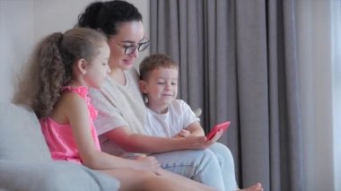 Mutlu aile, anne ve tatlı çocuklar, anne evde çocuklarla oynuyor rahatlıyor akıllı bir telefon kullanıyor kanepeye oturuyor kız ve oğul gülüyor, komik videolar izliyor, eğleniyor, aile hayatının tadını çıkarıyor.