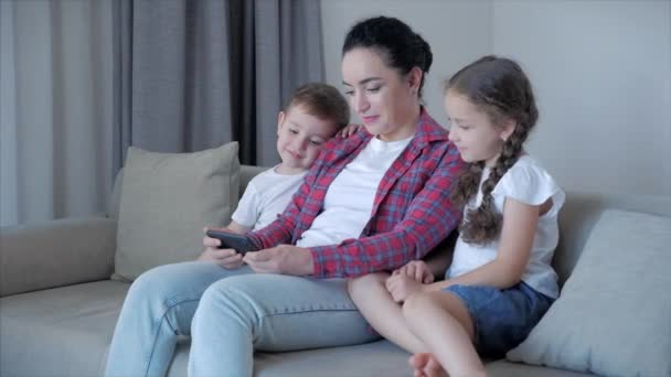 Família feliz, mamãe e filhinhos bonitos, mãe brincando com as crianças em casa relaxante usar um smartphone abraçando sentar no sofá filha e filho rir, assistir vídeos engraçados, se divertir, desfrutar de momentos da vida familiar — Vídeo de Stock