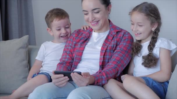 Щаслива сім'я, мама і милі маленькі діти дивляться телефон, мама грає з дітьми вдома, розслабляється за допомогою смартфона, сидить на дивані, сміється, розважається, насолоджується моментами сімейного життя — стокове відео