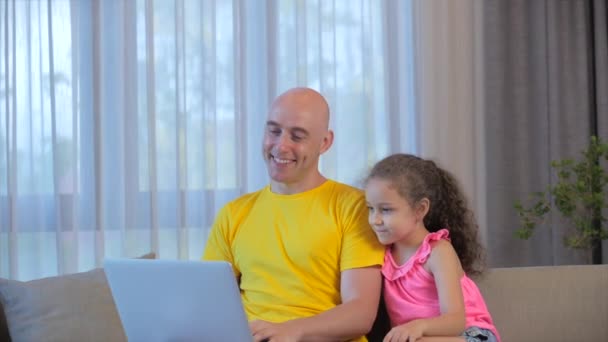 Jovem homem sério freelancer trabalhando em um laptop sentado em um sofá em casa, usando um computador para estudar o trabalho on-line em casa, sua filha bebê se aproxima dele e abraça seu pai cansado do trabalho. — Vídeo de Stock