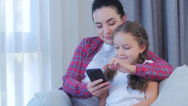 행복 한 엄마와 귀여운 딸, 집에서 쉬고 있는 아이들 과놀고 있는 엄마는 스마트폰을 사용하여 소파 딸에게 껴안고, 재밌는 비디오를 보고, 즐겁게 가족 생활을 즐긴다 — 비디오