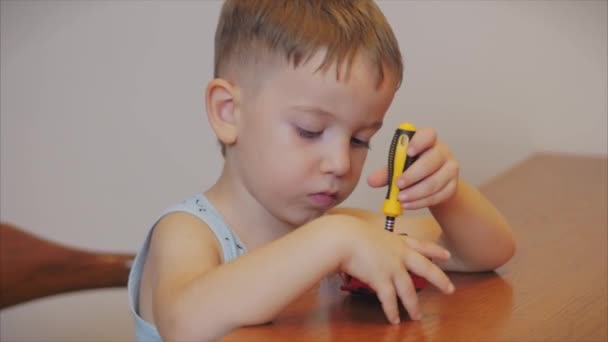 Skjuta ett porträtt av en söt förskola pojke, reparerar barnet sin favorit röd leksak bil för barn med en skruvmejsel, skruvar en skruv, utför en riktig arbetar sitter vid ett bord hemma. — Stockvideo