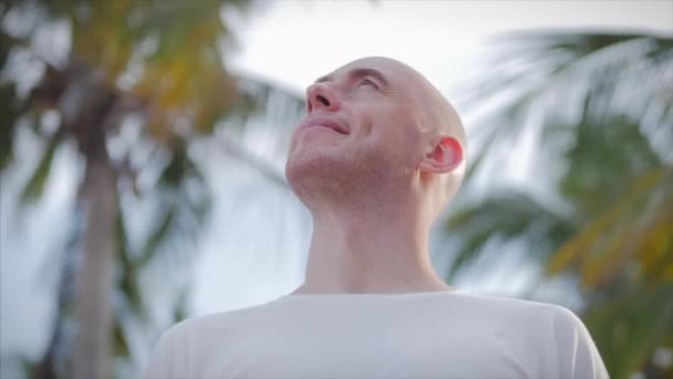 Egy fiatal férfi portréja, aki az utcára néz a pálmafák és a napsütéses időjárás hátterében, egy férfi, aki felnéz az égre és boldogan mosolyog.. — Stock videók