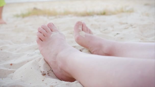 Närbild kvinnliga nakna fötter på en sandstrand i soligt soligt soligt väder ansiktslös barfota kvinna koppla av på stranden utomhus, mot bakgrund av ett barn som leker på stranden med sand och havsvatten. — Stockvideo