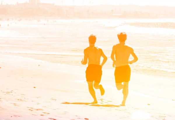 Two men running on sand near sea, ocean, water during sunset, people running on beach, silhouettes of runners