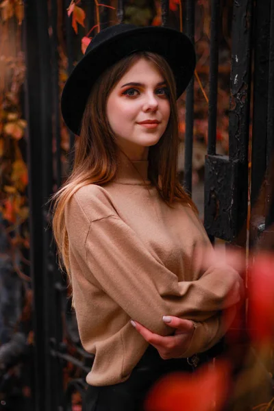 Jesienna dziewczyna stoi przy ogrodzeniu z czerwonym bluszczem. Sweter, kapelusz i... — Zdjęcie stockowe