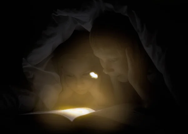 Hermana y hermano están leyendo un libro bajo una manta con linterna. bastante joven chico y encantadora chica tener divertido en niños habitación . — Foto de Stock