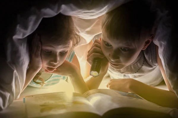 儿童睡前。姐姐和弟弟正在阅读一本书下毛毯，手电筒。年轻漂亮的男孩和可爱的女孩在儿童房开心. — 图库照片