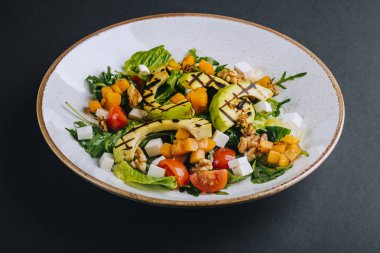 Balkabaklı sıcak salata, ızgara avokado, vişneli domates ve siyah arka planda beyaz peynir.