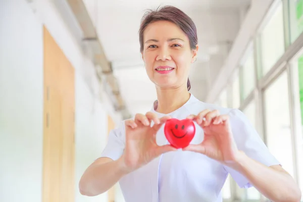 Smiling female nurse holding red smile heart in her hands. Red heart Shape representing high quality service mind to patient. Professional, Specialist  in white uniform and Valentine concept