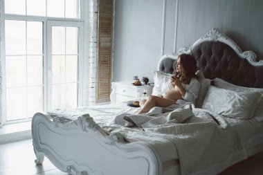 Kar-beyaz bir yatak odasında hamile bir kızla yatakta kahvaltı