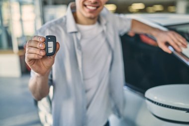 Smiling man holding a key behind a car in showroom. Business con