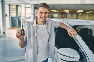 Smiling man holding a key behind a car in showroom. Business con