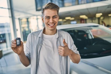 Smiling man holding a key behind a car in showroom. Business con