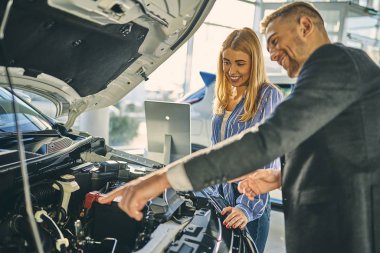 Smiling sale manager showing a car engine. Cheerful girl choosin