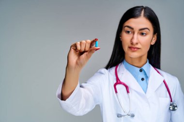 Koyu saçlı, kırmızı tırnaklı bir kadın parmaklarında mavi bir ilaç tutuyor.