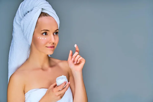 Calm woman in bath towel with towel on her hair with cream on cheek and finger with jar of cream in her hand
