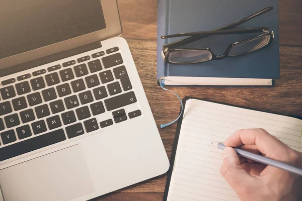 Top view of the hand of a male person taking notes on a notepad beneath a notebook computer on a wooden desktop — Stock Photo, Image