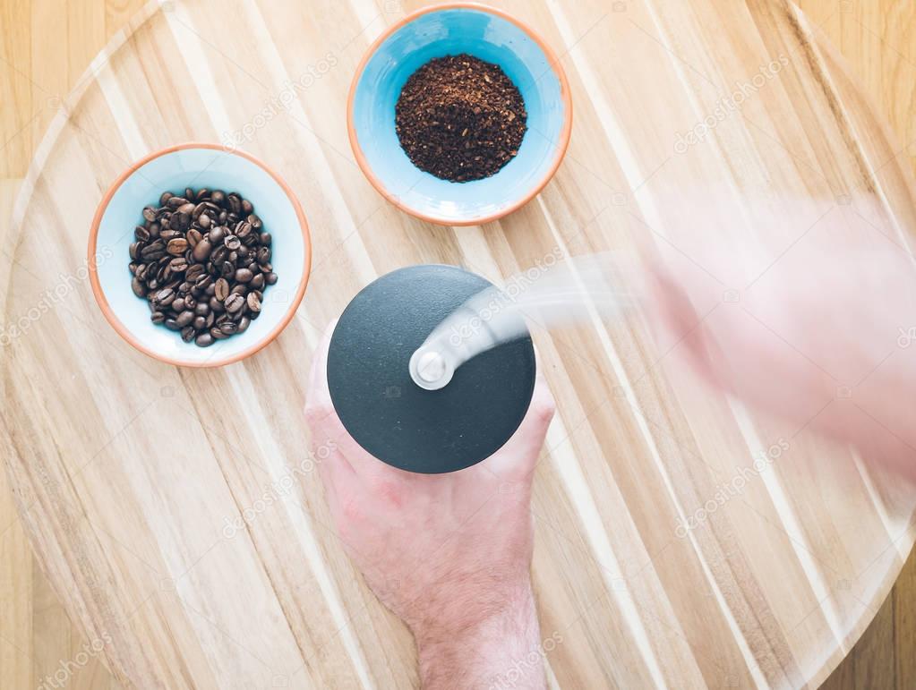top view of male hands operating a manual coffee grinder and two bowls with coffee beans and ground coffee on wooden surface