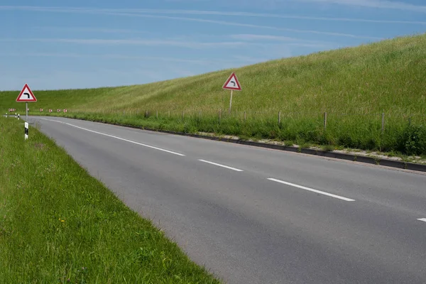 Señal de advertencia que indica curva izquierda aguda en ambos lados del camino vacío detrás de un dique cubierto con hierba verde exuberante —  Fotos de Stock