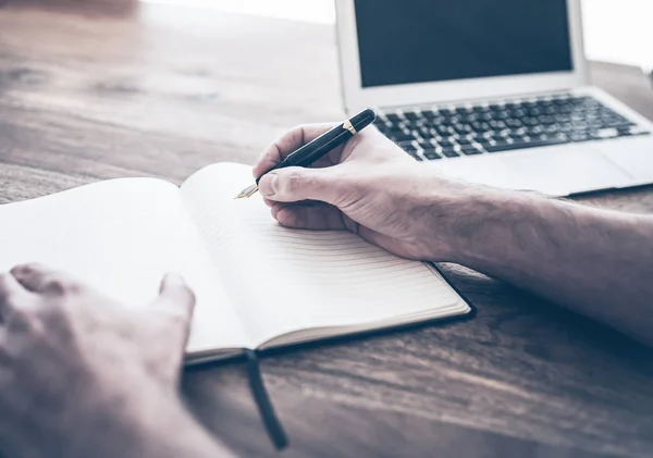 Manlig person skriver i anteckningsboken förutom bärbar dator på rustika trä skrivbord — Stockfoto