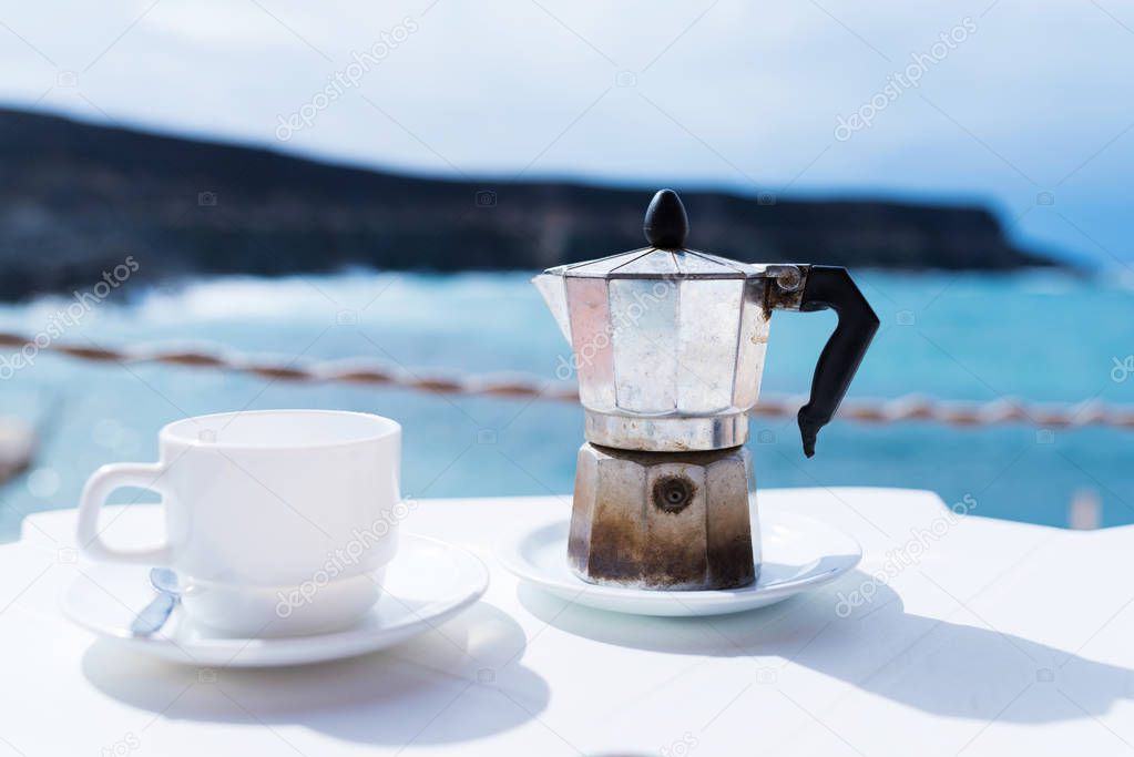 moka pot espresso maker and cup of coffee on table with coastline and ocean in background