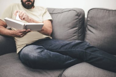 Sakallı, beyaz bir adam kanepeye uzanmış kitap okuyor.