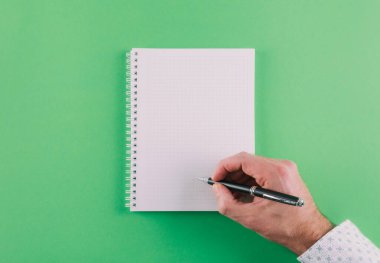 top view of hand holding ballpoint pen against spiral notepad on green background