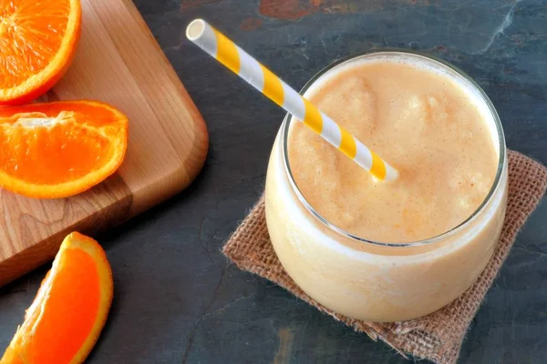 Healthy orange smoothie in a glass with striped straw and fresh fruit slices, downward view on slate