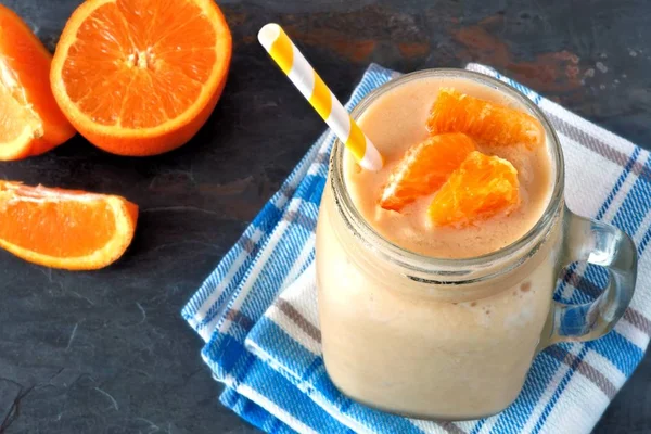 Orange fruit smoothie in a mason jar glass resting on checked cloth with fresh orange slices over a slate background