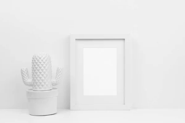 Mock up white frame and cactus decor on a shelf or desk. White color scheme. Portrait frame orientation.