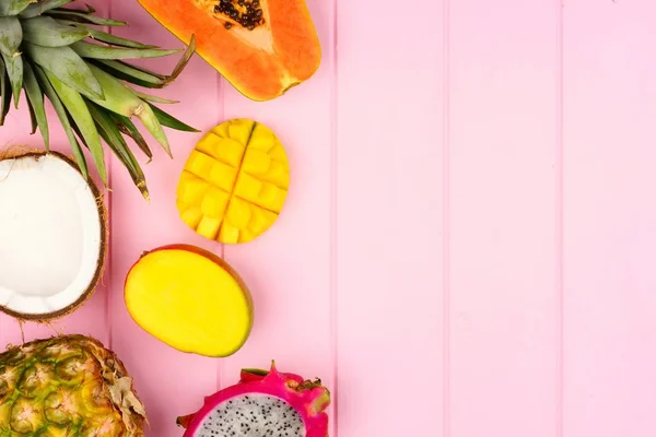 Tropical fruit side border with pineapple, dragon fruit, papaya, coconut and mango on a pink wood background