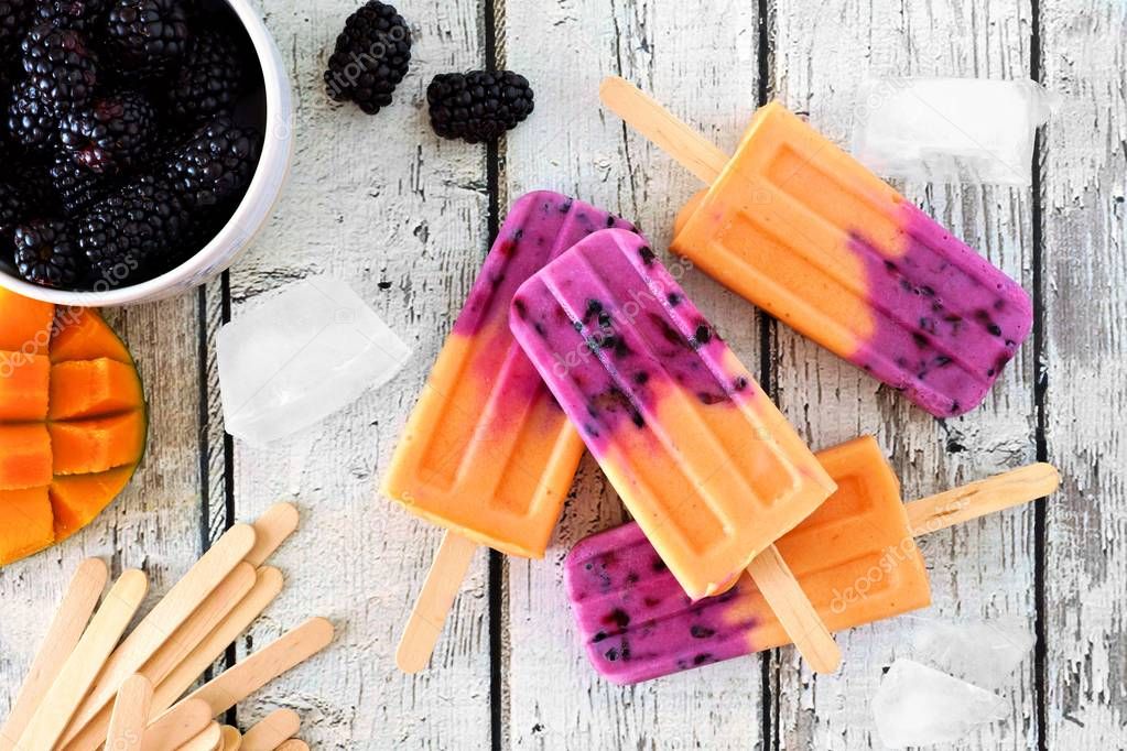 Group of homemade mango blackberry popsicles, top view on a rustic white wood background