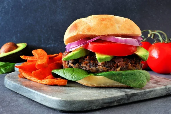 Vegetarian bean burger with avocado, spinach and sweet potato fries against a dark background