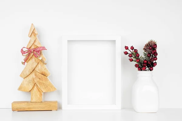 Mock up white frame with wooden Christmas tree and branch decorations on a shelf. Portrait frame against a white wall.