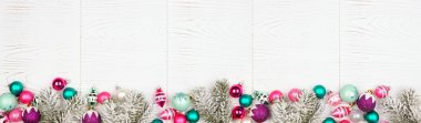 Christmas border banner with pink, purple and teal decorations and tree branches. Overhead view on a white wood background.