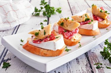 Peynirli, salamlı ve enginarlı crostini aperatifleri. Beyaz ahşap arka planda servis tabağına yakın dur. Parti yemeği konsepti.