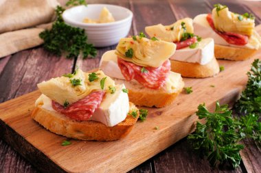 Peynirli, salamlı ve enginarlı crostini aperatifleri. Bir servis tahtasına, tahta bir arka plana yakın dur. Parti yemeği konsepti.