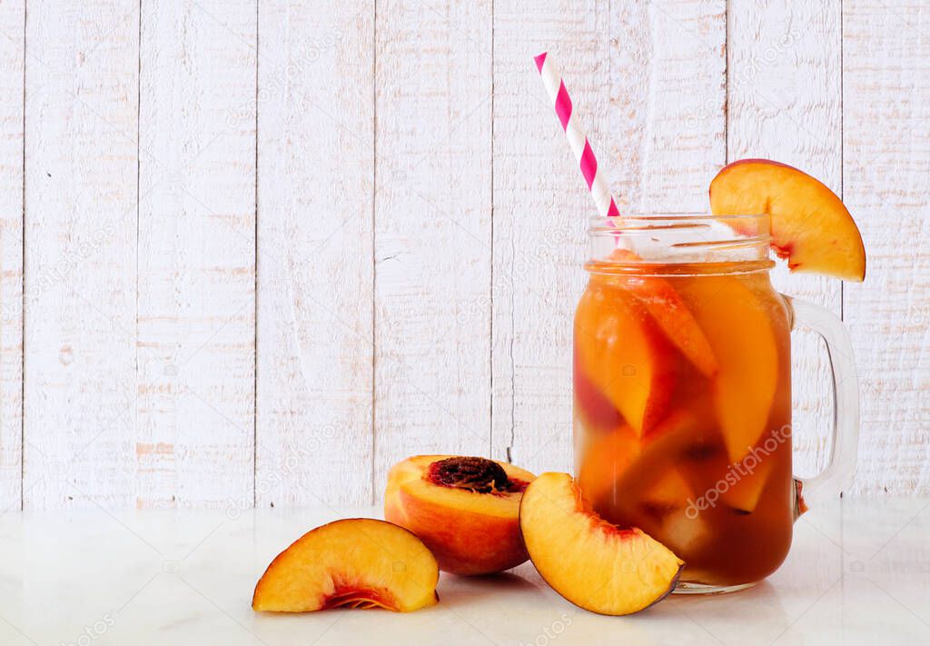Refreshing peach iced tea in a mason jar glass. Side view with cut fruit against a white wood background.