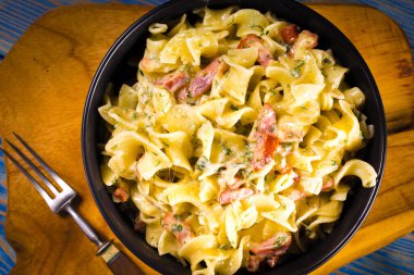 Ahşap bir masada siyah bir kasede sosisli ve peynirli makarna..