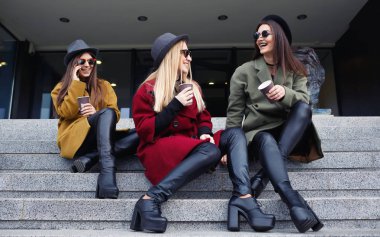 Şık kız trendy kat, eşarplar, şapka ve deri ayakkabı mutfak için merdivenlerde alışılmadık makyaj ile kahve içmek