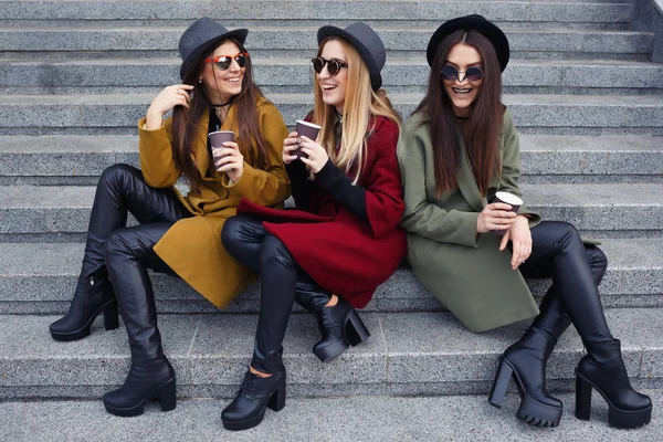 Stylish girls with unusual makeup in trendy coats, scarves, hats and leather shoes on the stairs to the galley drink coffee
