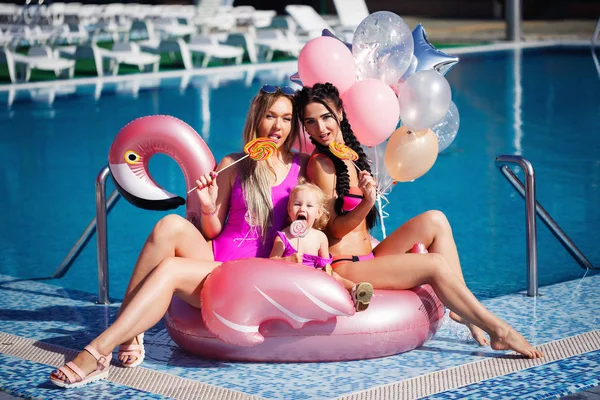 İki kadın bir bebekle şişme flamingo havuz etrafında oturan ve gülümseyen parlak mayolar