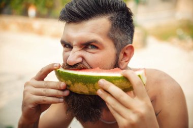 Bir adam şık bir saç modeli, uzun sakallı ve büyük bir zevk ile dövme karpuz yiyor