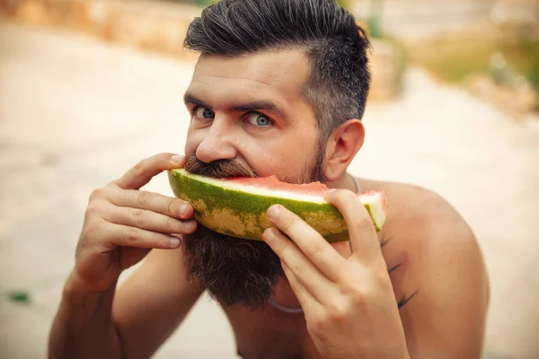 Un homme avec une coiffure élégante, une longue barbe et un tatouage avec un grand plaisir mange pastèque — Photo