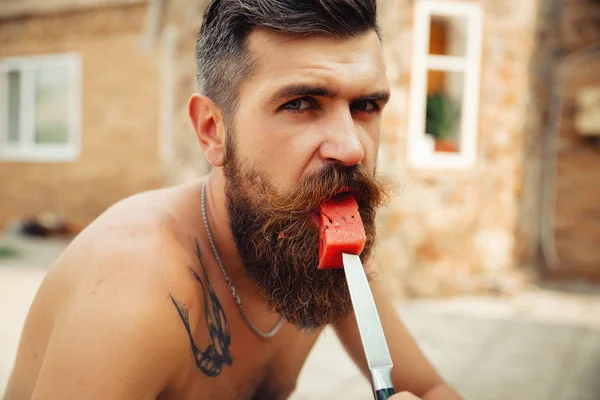 Bir adam şık bir saç modeli, uzun sakallı ve büyük bir zevk ile dövme karpuz yiyor