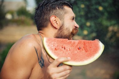 Bir adam şık bir saç modeli, uzun sakallı ve büyük bir zevk ile dövme karpuz yiyor