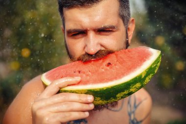 Islak, şık, sakallı, uzun bıyıklı esmer adam iştah açıcı bir şekilde sulu bir karpuzu yağmurda bir meyve bahçesinin arka planında yiyor.